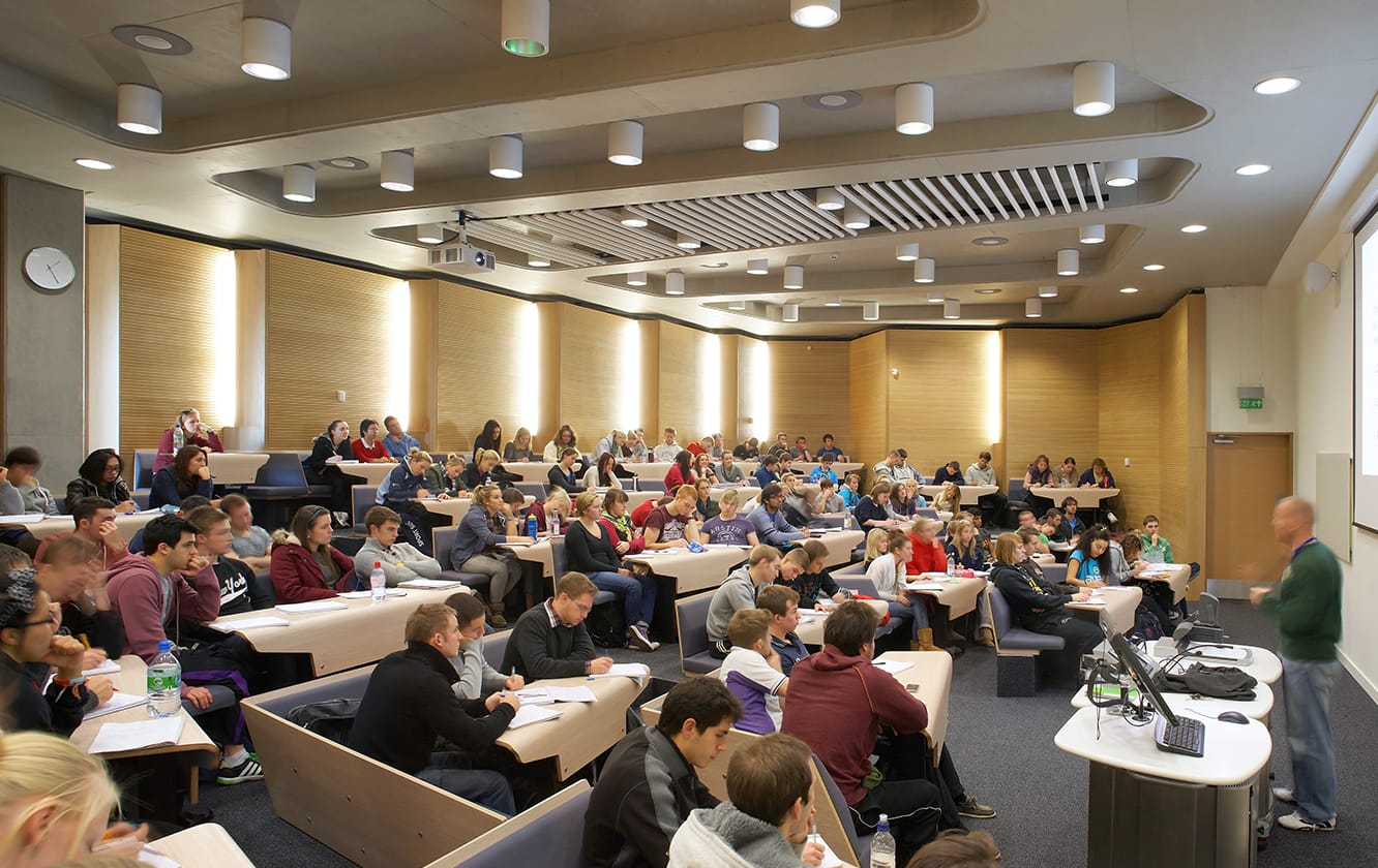 A lecture hall with blue lecture hall seating and desks filled with students