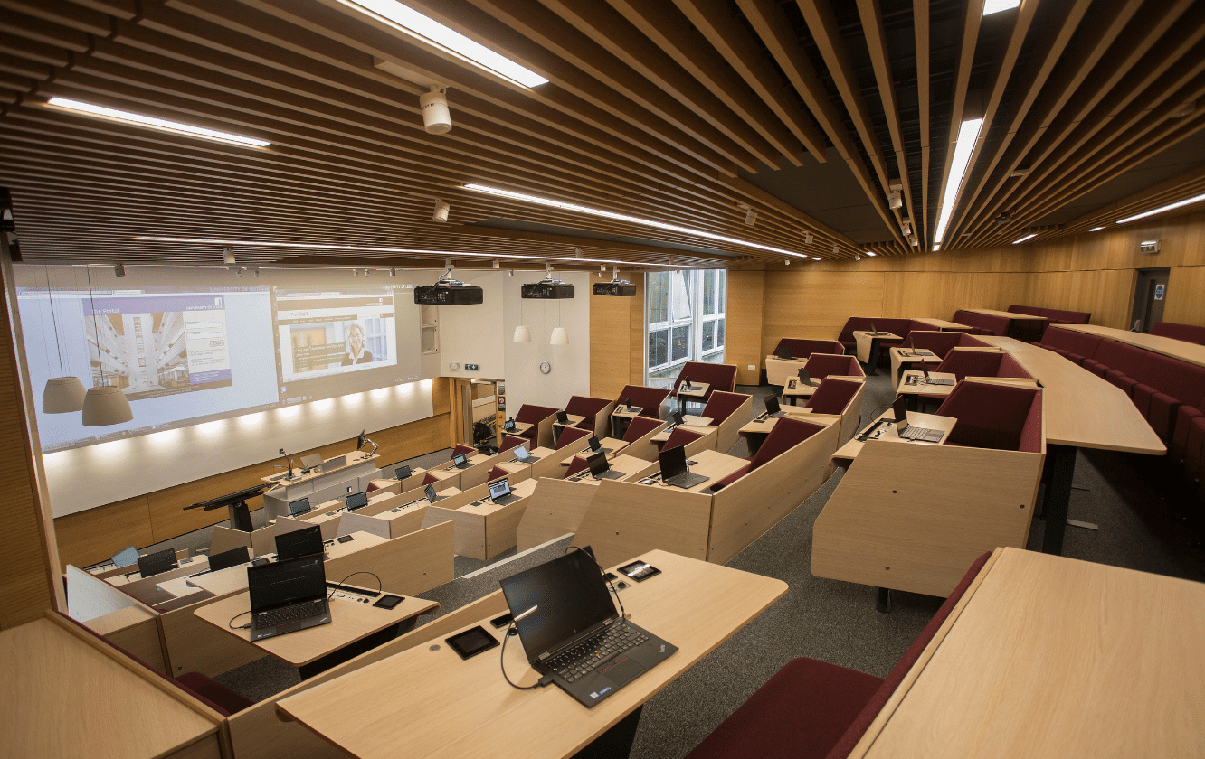A lecture hall with wooden desks, lecture hall seating and a projection screen.