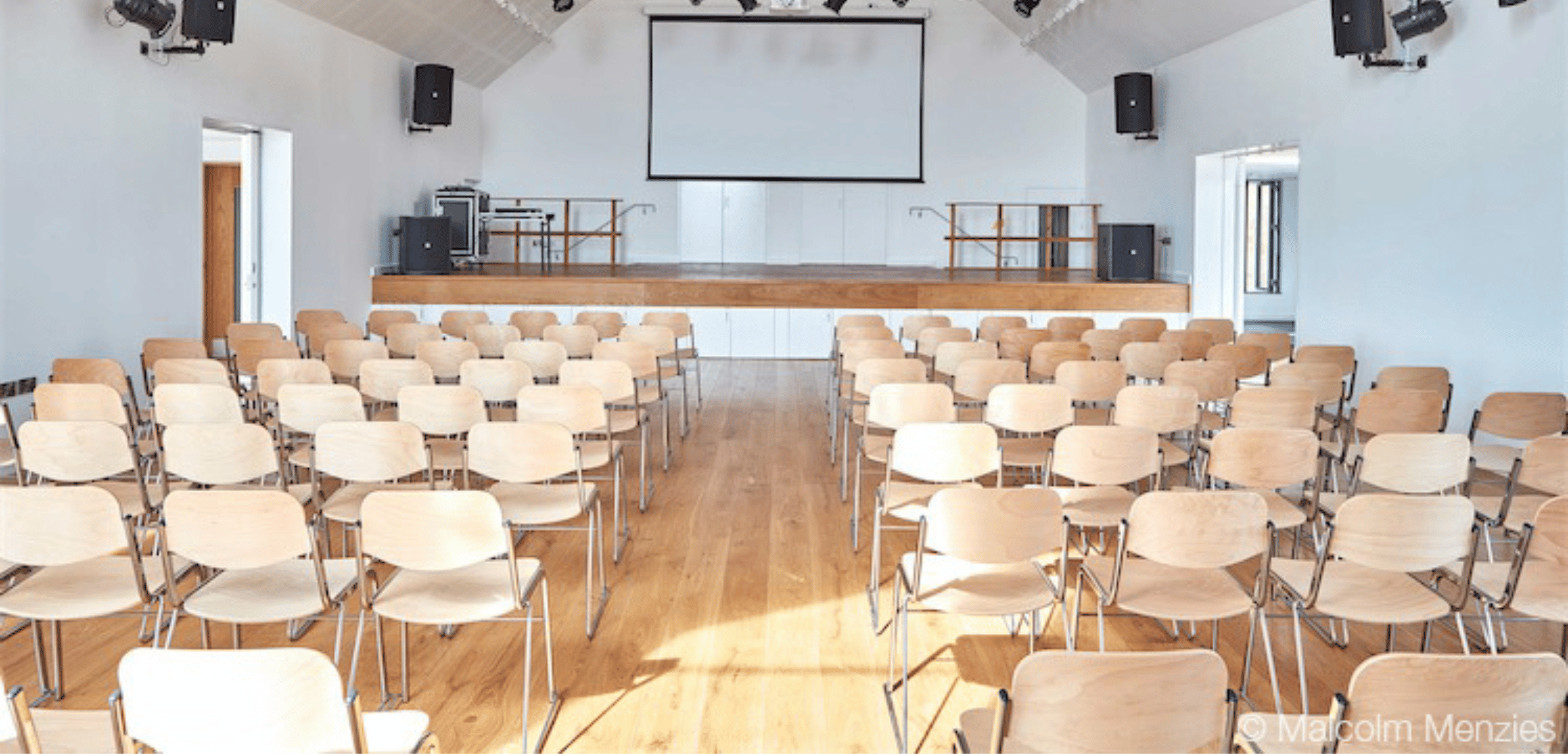 A large room with wooden community seating and a projector screen.